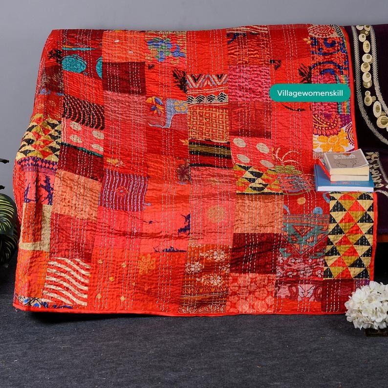 May include: A red patchwork quilt with a variety of colorful patterns and textures. The quilt is made of recycled saris and is hand-stitched. The quilt is folded and displayed on a couch with a book and flowers in the background. The text "Villagewomenskill" is visible in the upper right corner of the image.