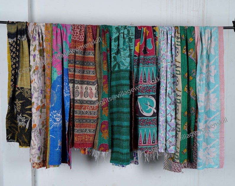 May include: A collection of colorful kantha quilts, also known as kantha stitch quilts, hanging on a white wall. The quilts are made of recycled saris and feature a variety of patterns and colors, including blue, green, pink, and yellow.  The quilts are all hand-stitched and have a unique, vintage look.