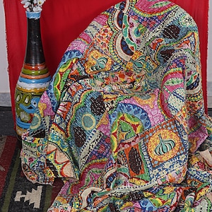 May include: A colourful kantha quilt with a patchwork design featuring various patterns and colours, including pink, blue, green, yellow, and brown. The quilt is made of cotton and is hand-stitched. It is folded and draped over a chair.