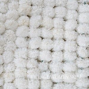 May include: A close-up of a wall of white paper pom-poms. The pom-poms are arranged in a random pattern and are all the same size. The text "VILLAGE WOMEN SKILL" is visible at the bottom of the image.