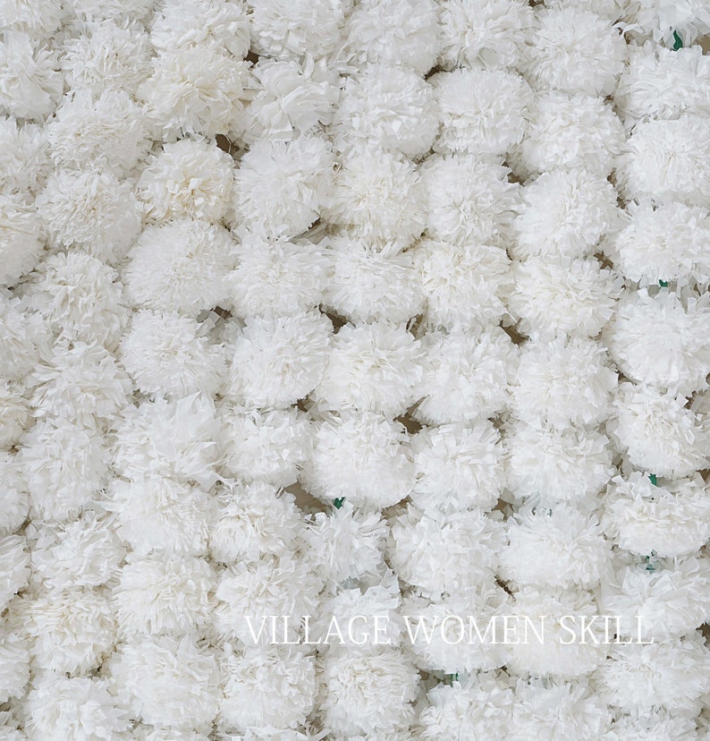 May include: A close-up of a wall of white paper pom-poms. The pom-poms are arranged in a random pattern and are all the same size. The text "VILLAGE WOMEN SKILL" is visible at the bottom of the image.