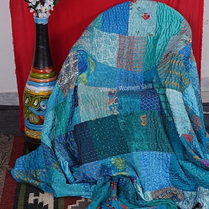 May include: A patchwork quilt made from repurposed saris in shades of blue, teal, and turquoise. The quilt is folded over a chair and has a tag that reads "Village Women Skill".