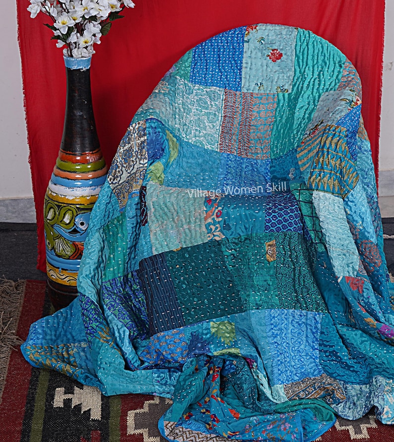 May include: A patchwork quilt made from repurposed saris in shades of blue, teal, and turquoise. The quilt is folded over a chair and has a tag that reads "Village Women Skill".