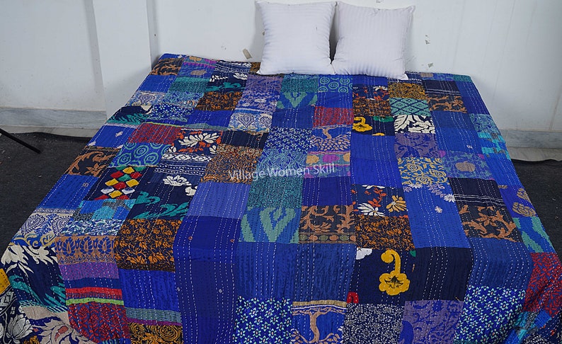 May include: A blue kantha quilt with a patchwork design made from various fabrics. The quilt is made up of squares of different colors and patterns, including floral, geometric, and paisley designs. The quilt is laid out on a bed with two white pillows.