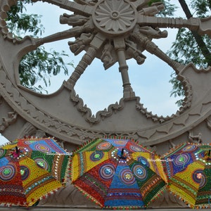May include: Three colorful, decorative umbrellas with intricate patterns and mirror accents are displayed. The umbrellas are open, showcasing a vibrant mix of colors including red, yellow, blue, and green. A large, carved stone wheel is in the background.