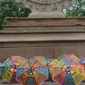 May include: A collection of colourful, patterned umbrellas with intricate embroidery and mirror embellishments. The umbrellas are arranged in a row on a stone surface in front of a stone structure.