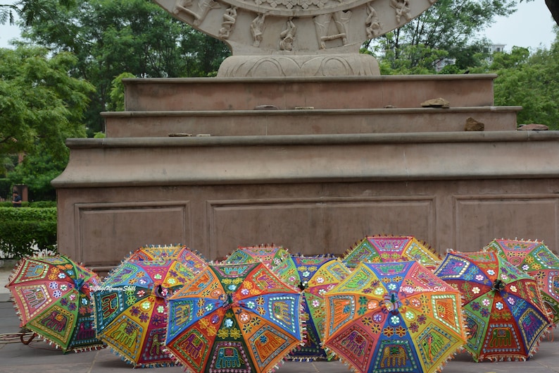 May include: A collection of colourful, patterned umbrellas with intricate embroidery and mirror embellishments. The umbrellas are arranged in a row on a stone surface in front of a stone structure.