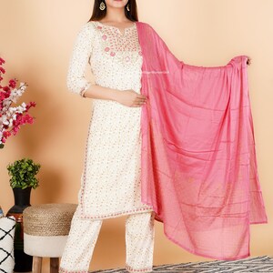 May include: A white cotton kurta with gold and pink embroidery, paired with white trousers and a pink dupatta. The kurta has a V-neckline and three-quarter sleeves. The dupatta has a gold border and is embroidered with small pink flowers.