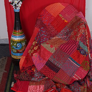 May include: A red patchwork quilt with intricate embroidery and a variety of patterns. The quilt is made of different fabrics and colors, including red, pink, orange, and blue. The quilt is folded and draped over a chair. The text "Village Women Skill" is visible on the quilt.