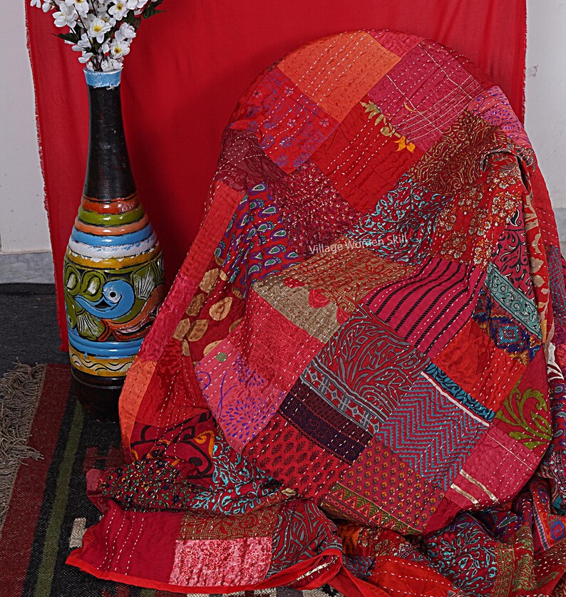 May include: A red patchwork quilt with intricate embroidery and a variety of patterns. The quilt is made of different fabrics and colors, including red, pink, orange, and blue. The quilt is folded and draped over a chair. The text "Village Women Skill" is visible on the quilt.