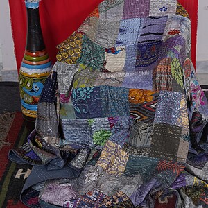 May include: A patchwork quilt made from a variety of colorful fabrics, featuring a variety of patterns and textures. The quilt is draped over a chair and has a gray background with a variety of colors, including purple, blue, green, and orange. The quilt is hand-stitched and has a unique, handmade look. The quilt is labeled "Village Women Skill".