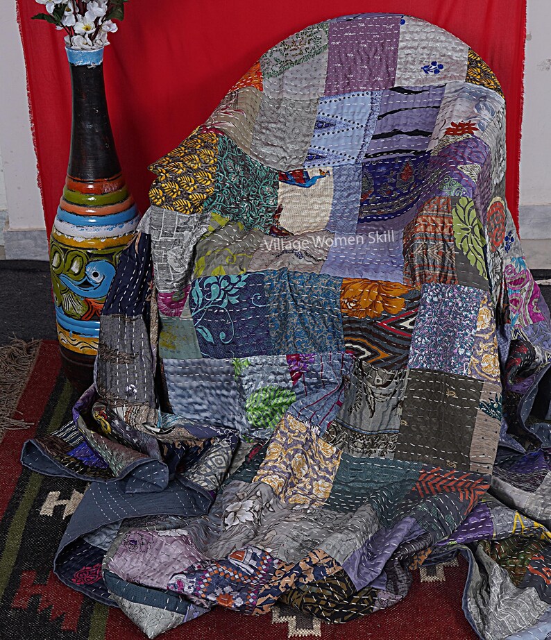 May include: A patchwork quilt made from a variety of colorful fabrics, featuring a variety of patterns and textures. The quilt is draped over a chair and has a gray background with a variety of colors, including purple, blue, green, and orange. The quilt is hand-stitched and has a unique, handmade look. The quilt is labeled "Village Women Skill".