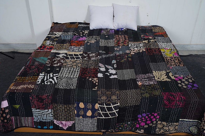 May include: A black kantha quilt with a patchwork design featuring various patterns and colors. The quilt is made of recycled saris and is hand-stitched. The quilt is laid out on a bed with two white pillows.