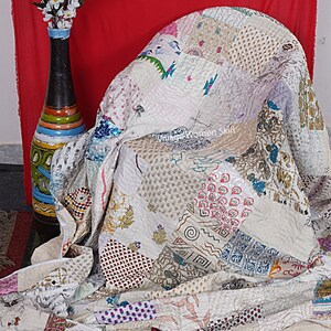 May include: A white patchwork quilt with a variety of colorful patterns and designs. The quilt is made of different fabrics and is hand-stitched. The quilt is draped over a chair and has a white background with a variety of colors and patterns.