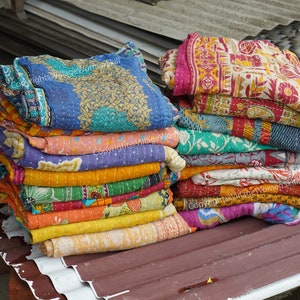 Puede incluir: Una pila de colchas kantha de colores, hechas con saris reciclados, con intrincados patrones y bordados. Las colchas est&aacute;n dobladas y apiladas sobre una superficie met&aacute;lica.