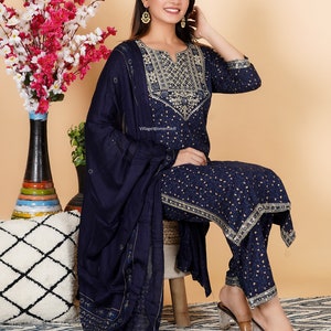 May include: A navy blue three-piece suit with gold embroidery. The suit includes a long-sleeved top, trousers, and a matching dupatta. The top has a V-neckline and a gold embroidered design on the front. The trousers are straight-leg and have a gold embroidered border. The dupatta is a sheer fabric with gold embroidery. The model is wearing the suit with clear heeled sandals.