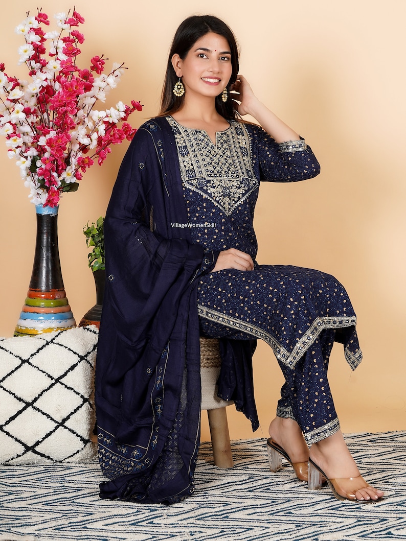 May include: A navy blue three-piece suit with gold embroidery. The suit includes a long-sleeved top, trousers, and a matching dupatta. The top has a V-neckline and a gold embroidered design on the front. The trousers are straight-leg and have a gold embroidered border. The dupatta is a sheer fabric with gold embroidery. The model is wearing the suit with clear heeled sandals.