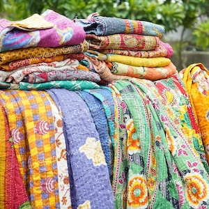 Op de afbeelding: Een verzameling kleurrijke, bedrukte quilts en dekens. De stoffen hebben verschillende ontwerpen, waaronder bloemen-, geometrische en patchworkpatronen. De quilts zijn gestapeld en gedrapeerd en tonen een reeks levendige kleuren en texturen.