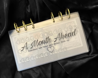 May include: A clear plastic binder with gold rings. The binder has a white background with black text that reads "A Month Ahead Remember the Goal and Never Give Up." There are small hearts printed on the background.