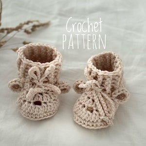 Könnte beinhalten: Ein Paar gehäkelte Babyschuhe in hellbeige. Die Schuhe haben ein schlichtes Design mit einem kleinen Gesicht auf der Vorderseite. Die Schuhe liegen auf einem weißen Stoffhintergrund. Häkelanleitung.