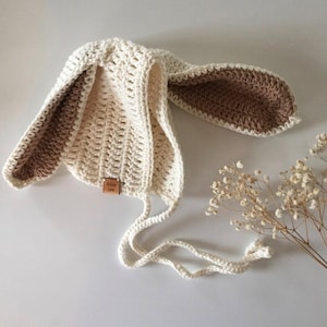 May include: A cream-colored crochet bonnet featuring long, floppy rabbit-style ears with brown inner detailing. The hat includes two chin ties and a small, rectangular tan leather tag on the front with the text Vintage Lane printed on it.
