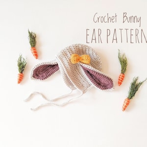 May include: Crochet bunny ear pattern with a gray and purple bunny hat with a yellow bow on top. The hat is laying on a white surface with small orange carrots with green tops.