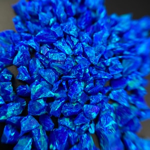 May include: A close-up of a pile of iridescent blue and green opal chips. The chips are irregular in shape and have a shimmering, reflective surface.