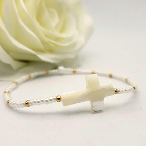 May include: A delicate beaded bracelet featuring a central white cross charm made of stone or shell material. The band consists of small white beads interspersed with tiny metallic gold-colored spacer beads, creating a simple and refined accessory.