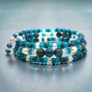 May include: A spiral wrap bracelet with blue, teal, and white beads. The bracelet features a mix of round and faceted beads, including pearls and crystal accents. The design is multi-layered, creating a textured and eye-catching accessory.