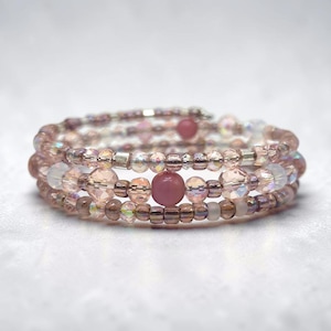 May include: A multi-strand wrap bracelet featuring a combination of translucent pink, iridescent clear, and opaque mauve beads. The design incorporates faceted crystal beads and smooth round beads arranged in a continuous spiral pattern.