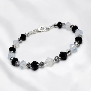 May include: A bracelet featuring alternating faceted black beads and iridescent white beads, accented with smaller dark gray crystal spacers. The piece is finished with a silver-toned metal lobster claw clasp and a short extender chain.