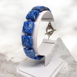 May include: A blue and white square bead bracelet with a silver clasp. The bracelet is displayed on a white jewelry stand. The beads have a marbled appearance, and the clasp is a toggle style.