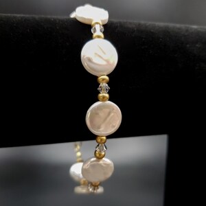 May include: A delicate bracelet featuring large, white, coin-shaped pearls interspersed with gold-colored beads and small, clear crystals. The bracelet is strung on a flexible cord, creating a simple yet elegant design. The background is black.