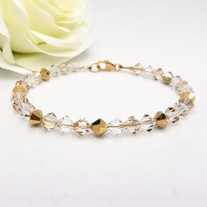 May include: A delicate gold bracelet featuring clear and gold-toned crystal beads. The bracelet has a gold clasp and is displayed on a white surface with a soft, white flower in the background. The crystals are faceted, catching the light.