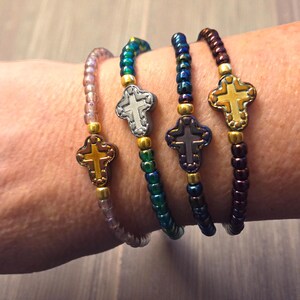 May include: Four beaded bracelets in shades of purple, green, blue, and brown, each featuring a cross charm. The bracelets are worn on a wrist, showcasing the different colors and designs. The crosses have gold and silver accents.