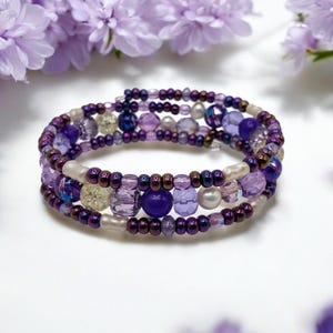 Puede incluir: Una pulsera de múltiples hilos con un diseño en espiral. La pulsera presenta una variedad de cuentas moradas, incluyendo pequeñas cuentas de semillas, cuentas facetadas más grandes y acentos de perlas. La paleta de colores incluye tonos de lavanda, amatista y morado intenso.