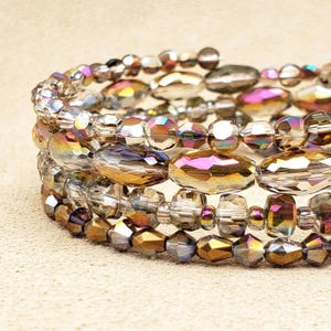 May include: Close-up of three beaded bracelets. The bracelets feature iridescent, faceted beads in shades of gold, purple, and clear. The beads are strung together, creating a layered effect. The bracelets are on a neutral background.