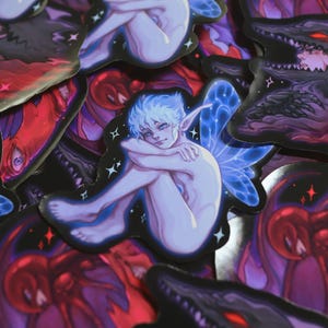 Peut inclure: Plusieurs autocollants sur le thème de la fantaisie. L'un d'eux représente une créature pâle aux cheveux et aux ailes bleus, entourée d'autres créatures sombres et fantastiques. Les autocollants ont une finition brillante et des couleurs variées, dont le bleu, le rouge et le violet.