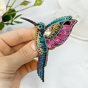 Może przedstawiać: Kolorowa broszka w kształcie kolibra z mieniącymi się niebieskimi, zielonymi i różowymi cekinami. Broszka ma duży kryształowy rhinestone w środku klatki piersiowej kolibra.