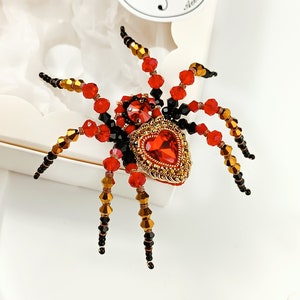 May include: A red and black beaded spider brooch with a gold heart-shaped centre. The heart is adorned with a red gemstone.