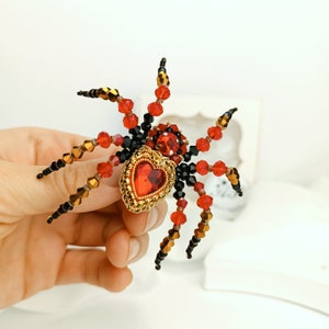 May include: A beaded spider brooch featuring a red heart-shaped gem at its centre. The spider is crafted with black, red, and gold beads.