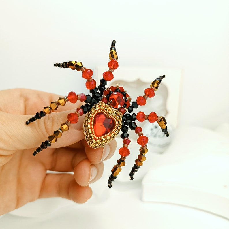 May include: A beaded spider brooch featuring a red heart-shaped gem at its centre. The spider is crafted with black, red, and gold beads.