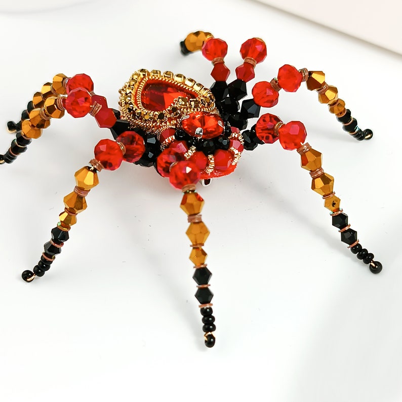 May include: A red and black beaded spider brooch with gold accents. The spider has a large red gemstone in the centre of its body.