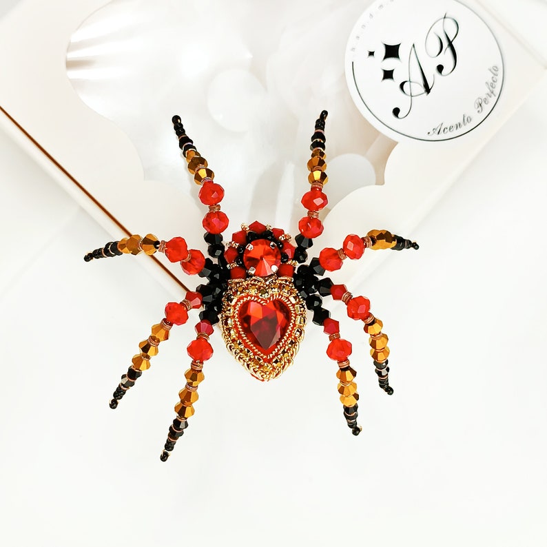 May include: A red and black beaded spider brooch with a large red heart-shaped rhinestone in the centre. The spider has eight legs and is made of small beads.