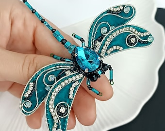 May include: A teal blue dragonfly brooch with a large, faceted, teal blue crystal in the center. The wings are embellished with silver wire, black beads, and clear rhinestones.