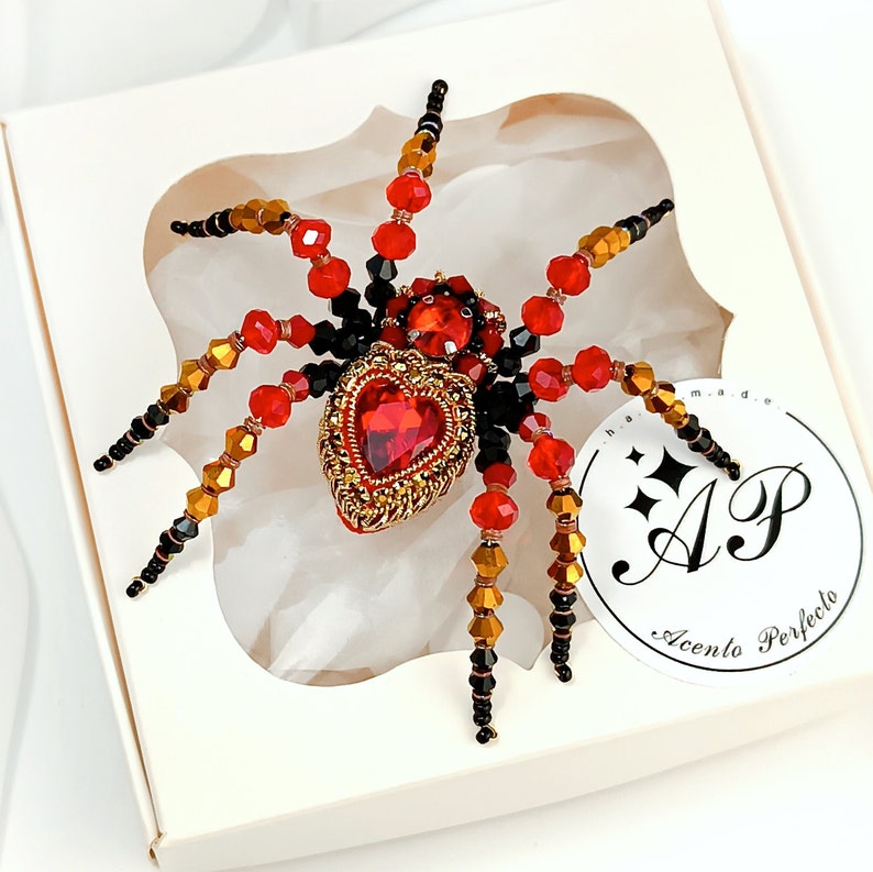 May include: A red and black beaded spider brooch with a large red heart-shaped gem in the centre. The brooch is on a white background with a logo that says "Accento Perfecto".