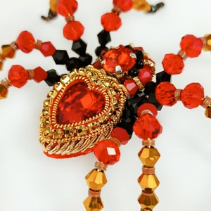 May include: A red and gold beaded spider brooch with a large red heart-shaped gem in the centre.