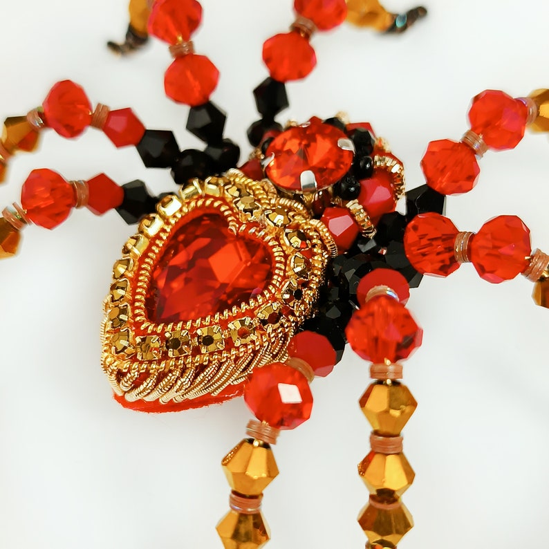 May include: A red and gold beaded spider brooch with a large red heart-shaped gem in the centre.