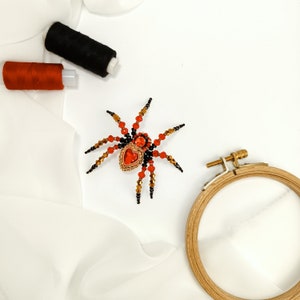May include: A beaded spider brooch with a red heart-shaped gem in the centre. The spider is made with black and red beads and has gold accents. The brooch is on a white fabric background.