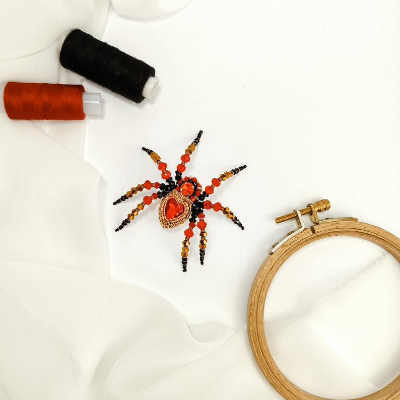 May include: A beaded spider brooch with a red heart-shaped gem in the centre. The spider is made with black and red beads and has gold accents. The brooch is on a white fabric background.